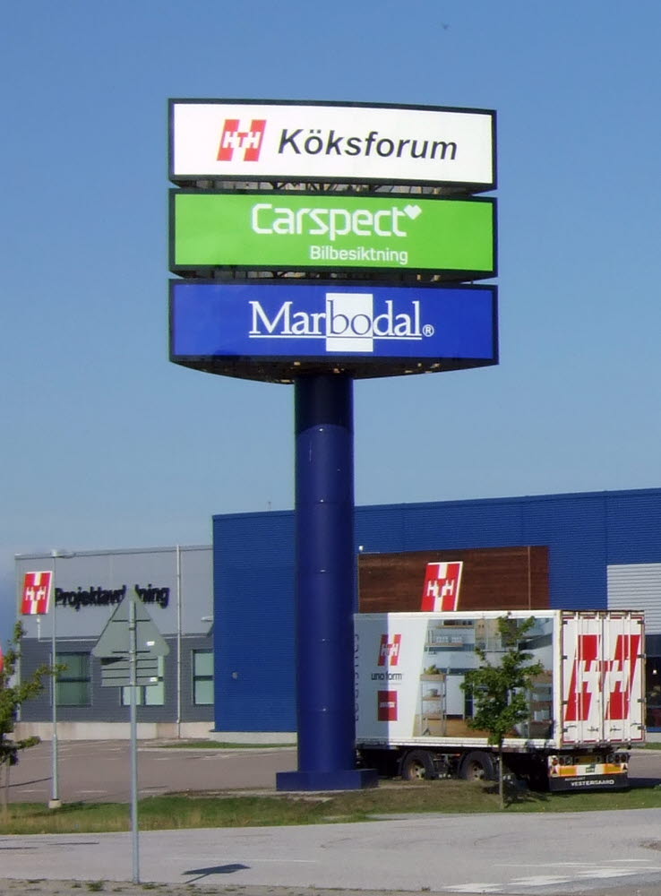 Foto av en hög stolpe med tre skyltar med texten Köksforum, Carspect Bilbesiktning och , Marbodal vid ett köpcentrum. 