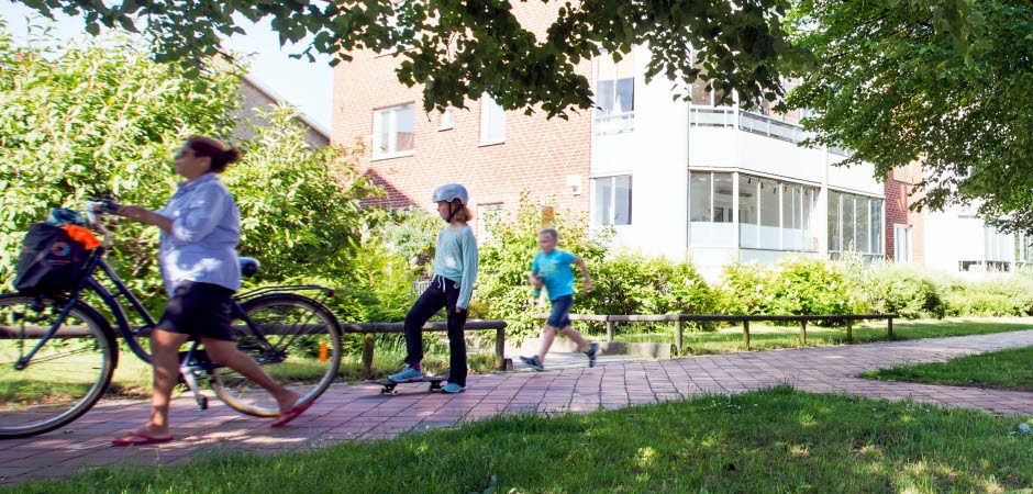 En cyklist, ett barn på skateboard och ett barn som springer på en gångbana med mycket grönska omkring.