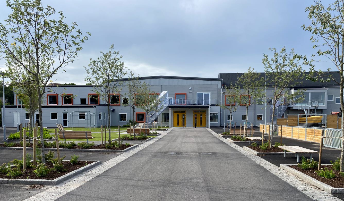 Foto på en grå tvåvåningsskola men skolgård i förgrunden.
