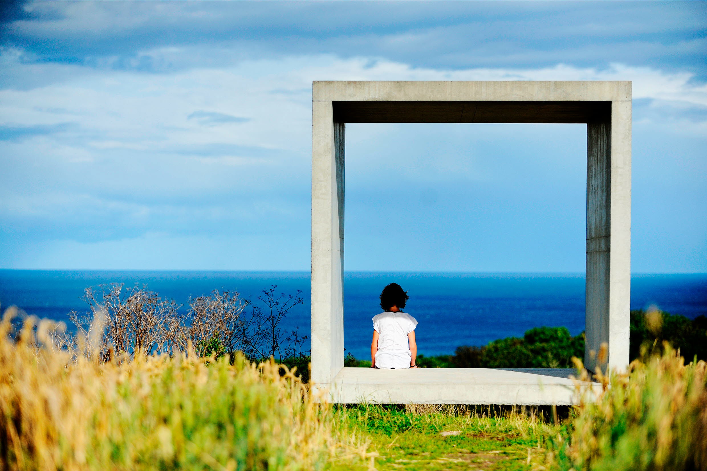 Foto på person som sitter i en ihålig kub av betong och tittar ut över havet.