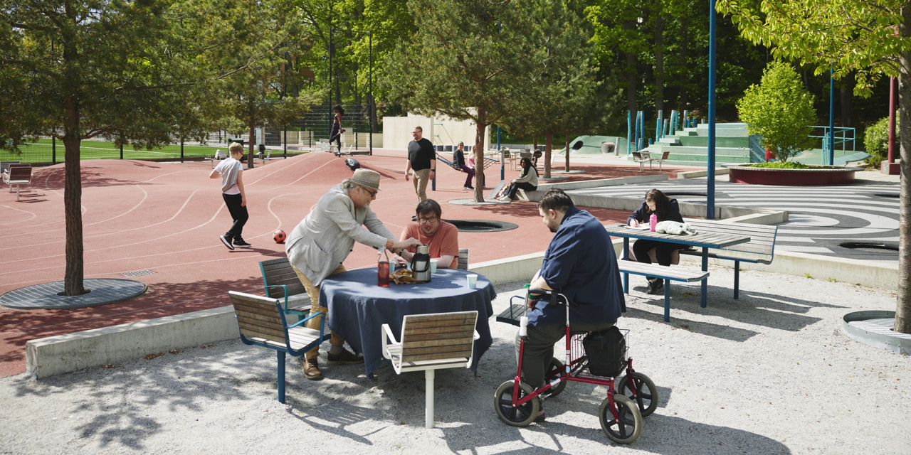 Foto på personer  som sitter vid bord och samtalar i en grön parkmiljö.