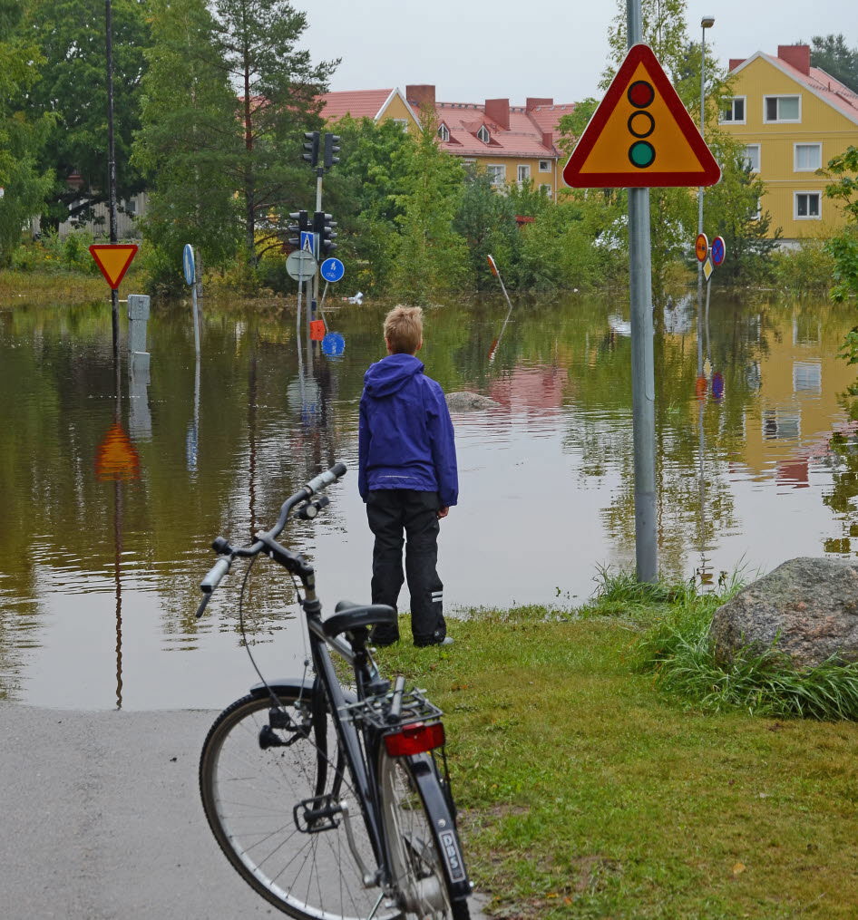 Ett barn har parkerat sin cykel vid en översvämmad vägkorsning. Det går inte att ta sig över till fots eller på cykel.