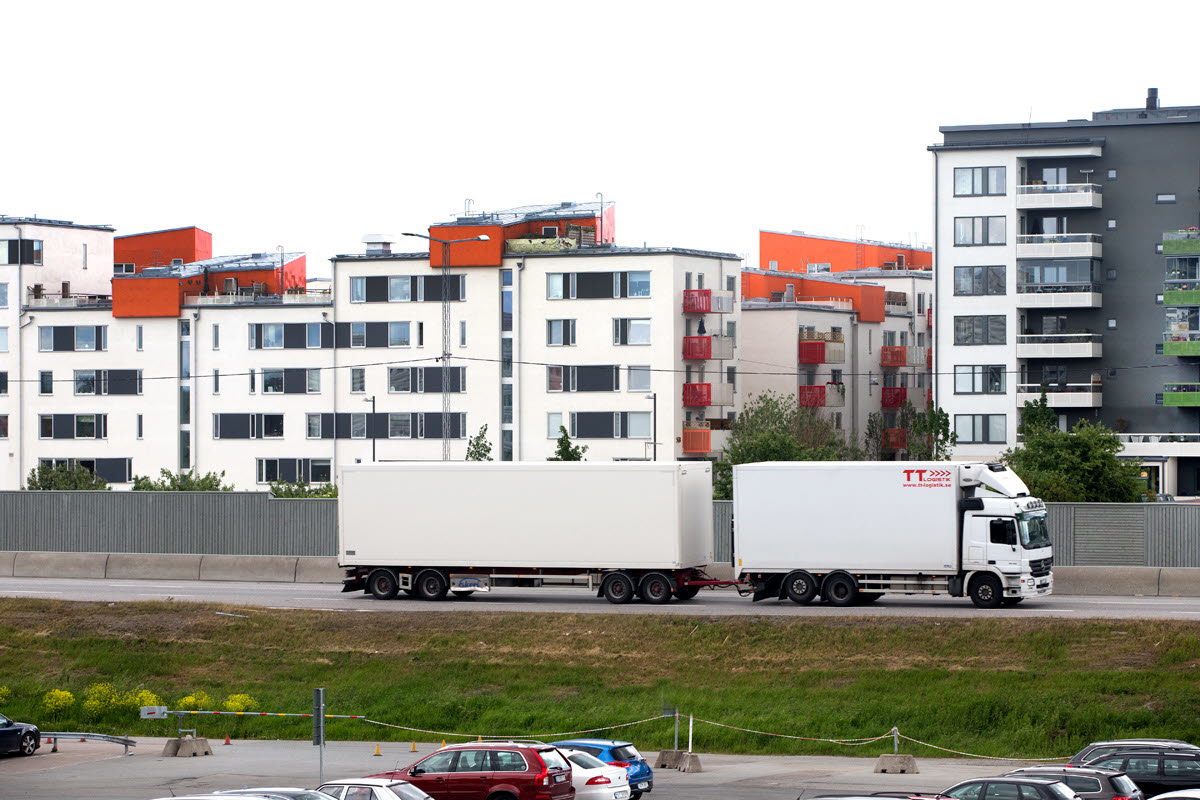 Foto av en lastbil som kör utanför ett bostadsområde med lägenheter och bullerplank.