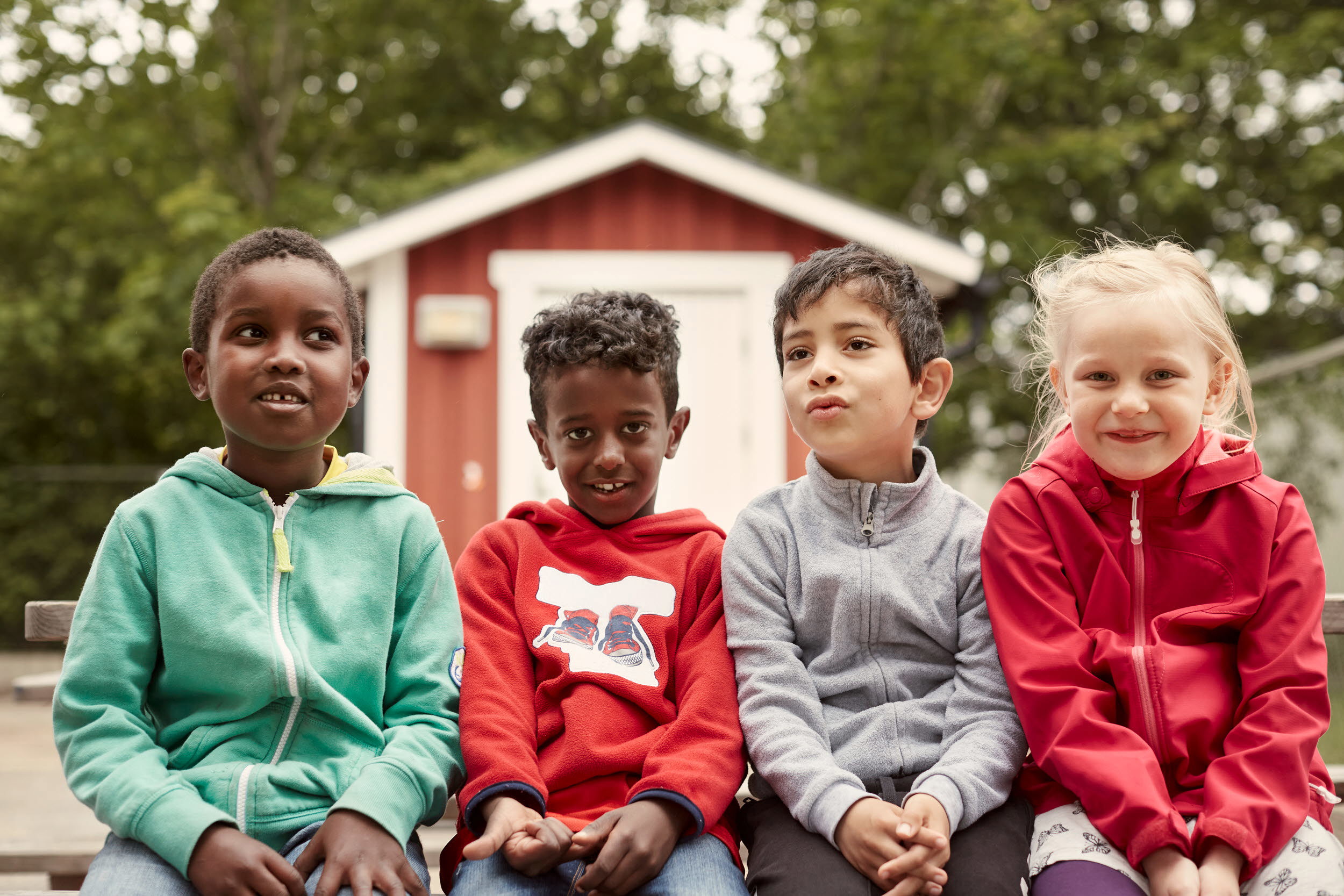 Foto med fyra barn som sitter på rad framför en faluröd bod.