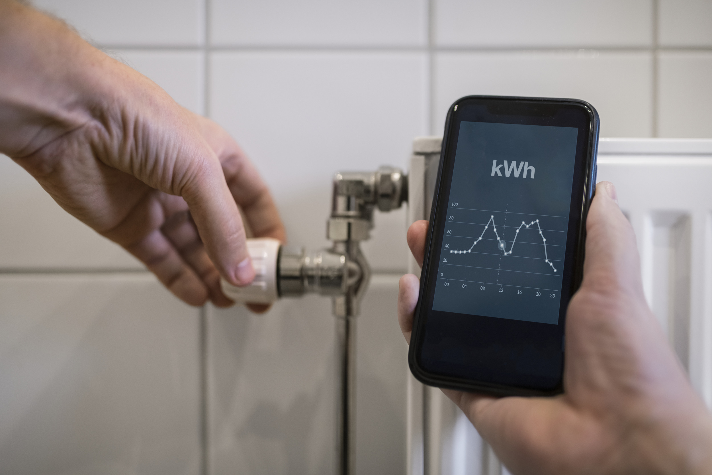 En person håller en mobiltelefon med kWh-mätning framför ett element. 