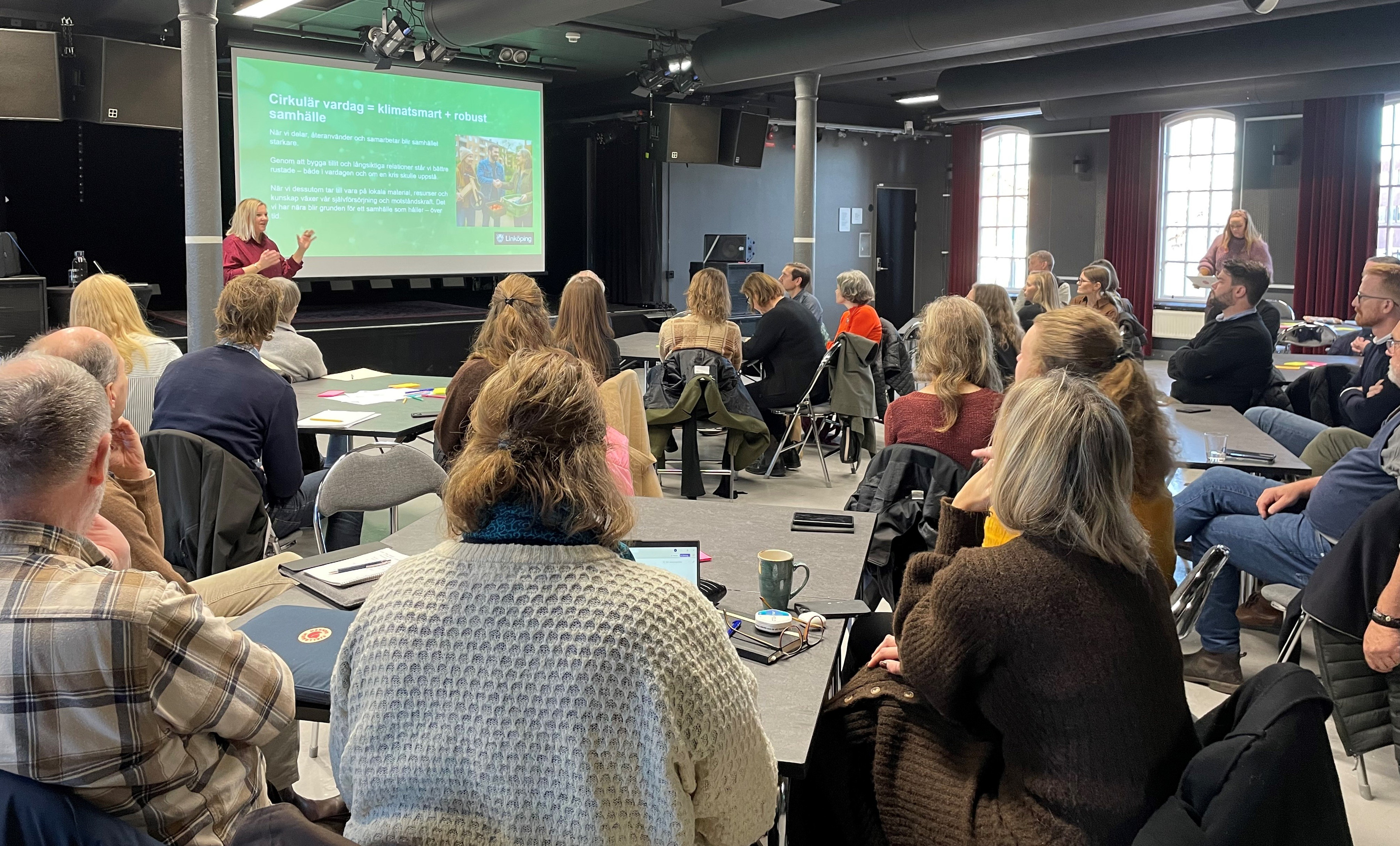 Flera människor sitter runt bord och tittar på en projektorduk.
