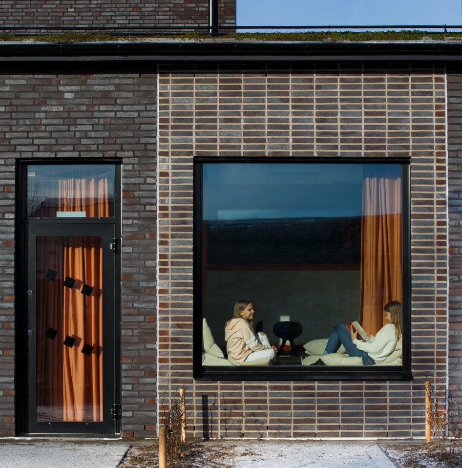 Foto på ett stort kvadratiskt fönster i en husfasad. Inne i fönsternischen sitter två flickor och läser.