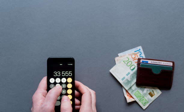 Hand som använder en miniräknare. Några sedlar och en plånbok ligger bredvid.