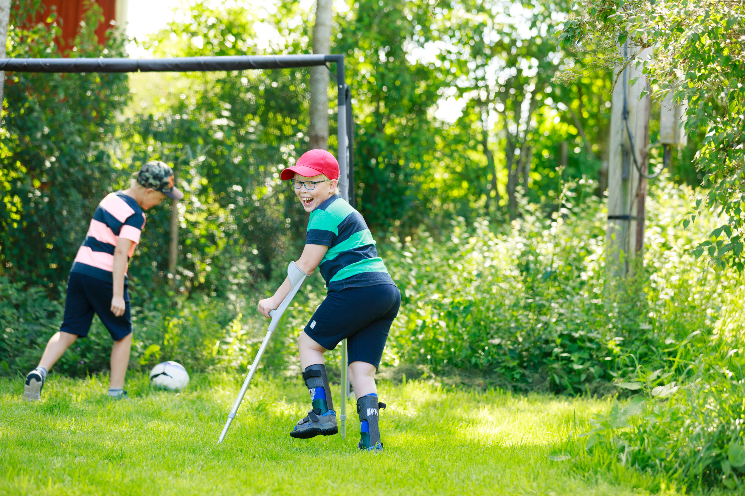 Fot på två barn spelar fotboll på en gräsmatta. Ett barn har kryckor och springer med dem som stöd.