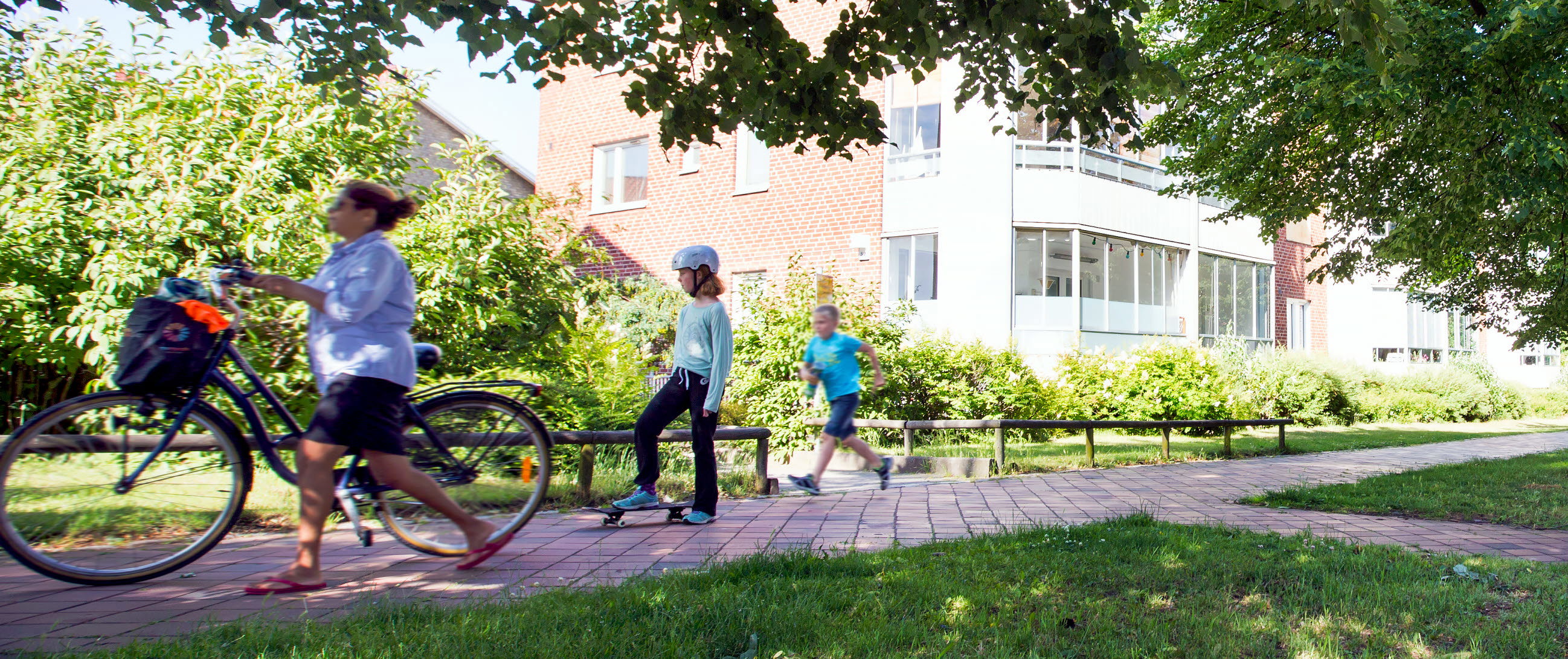 En cyklist, ett barn på skateboard och ett barn som springer på en gångbana med mycket grönska omkring.
