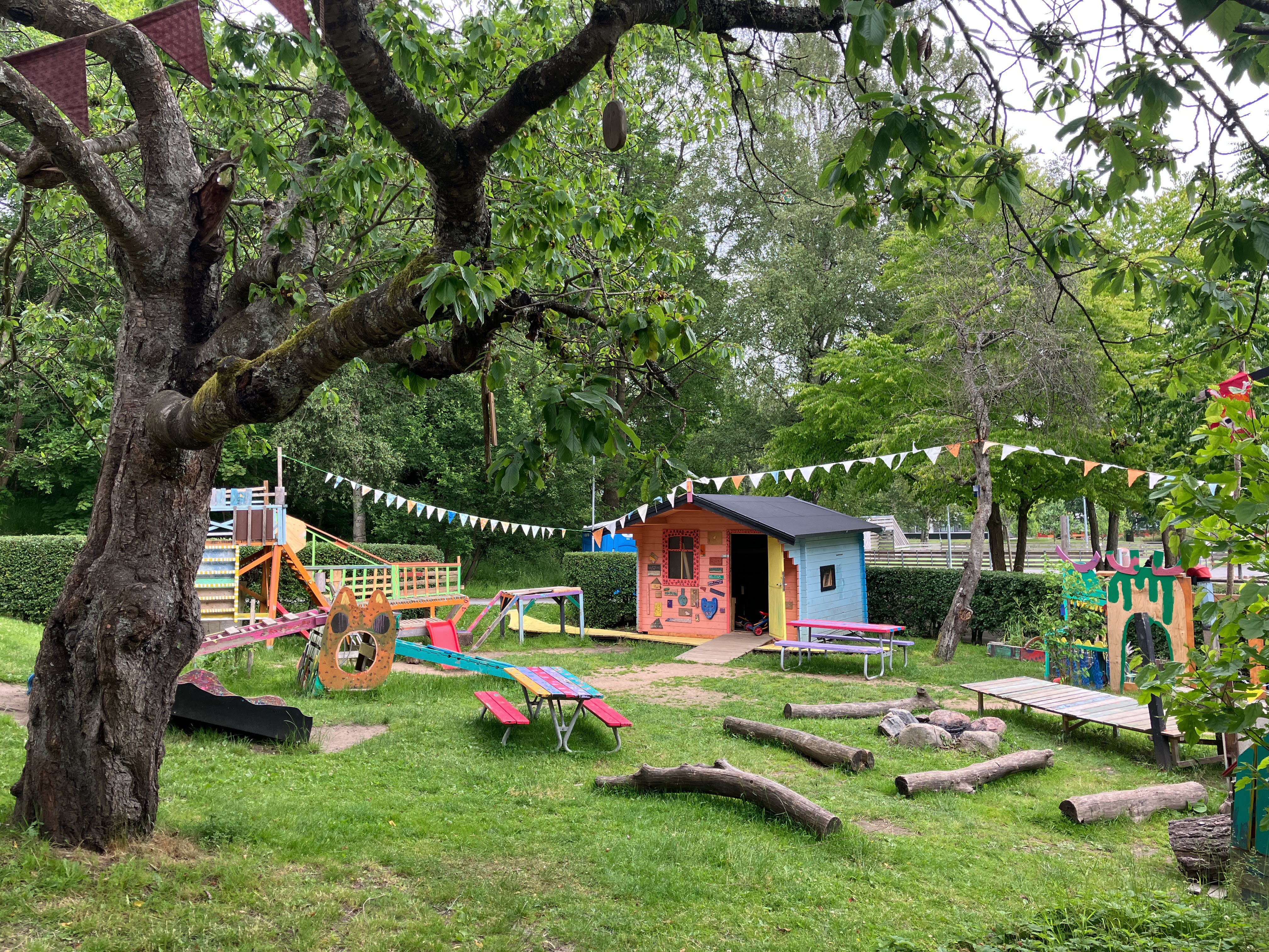 Bild på lekfull trädgård med färgglada vimplar, klätterträd, och bänkar. 