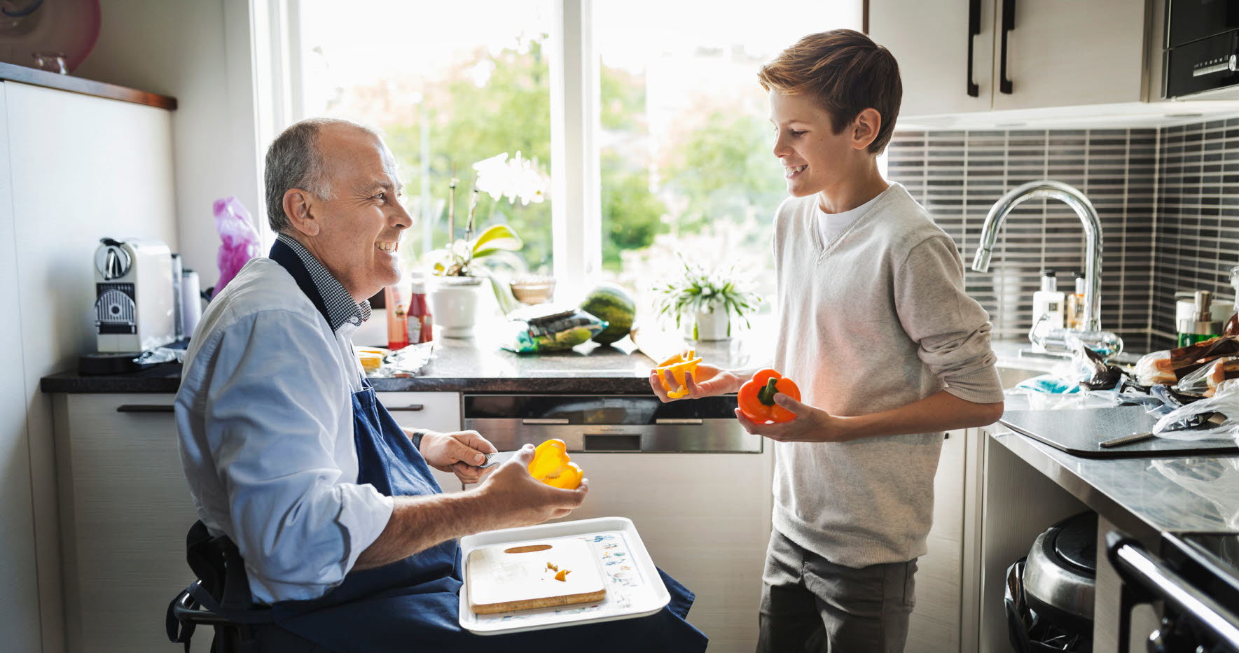 Pappa och son lagar mat i köket. Pappan använder rullstol. De är glada.