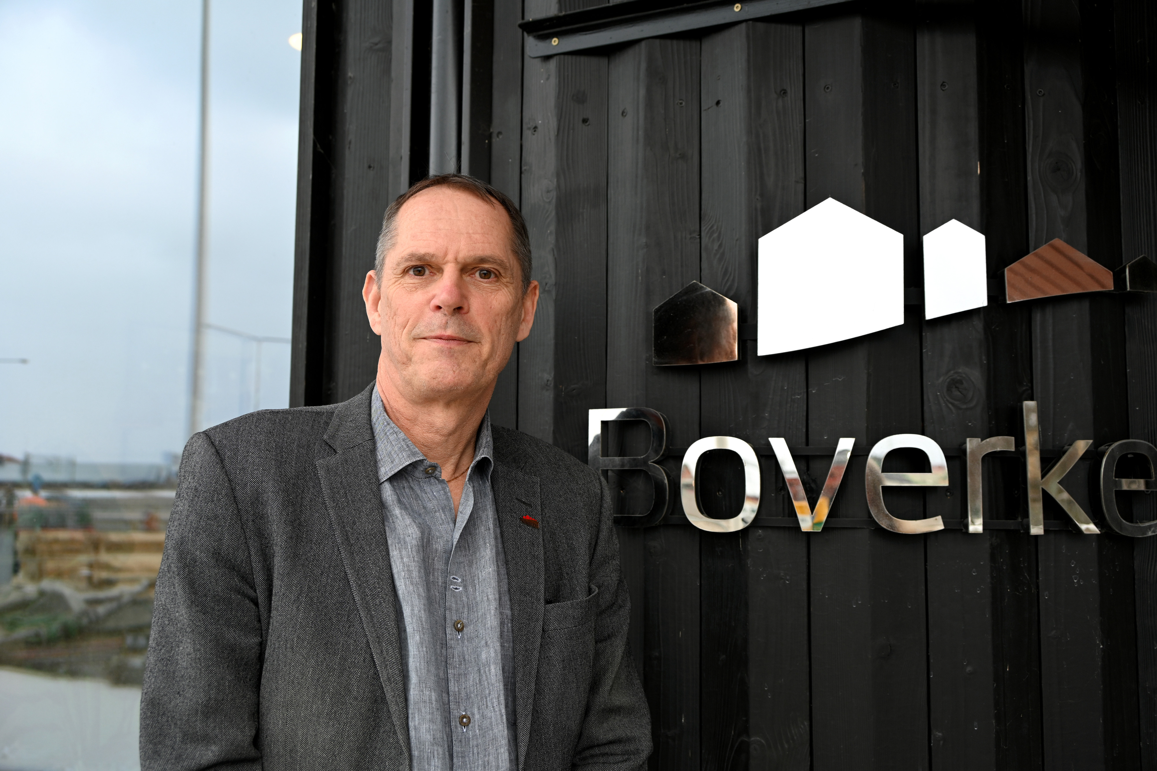 En man i brun kavaj framför en byggarbetsplats med en lyftkran. 