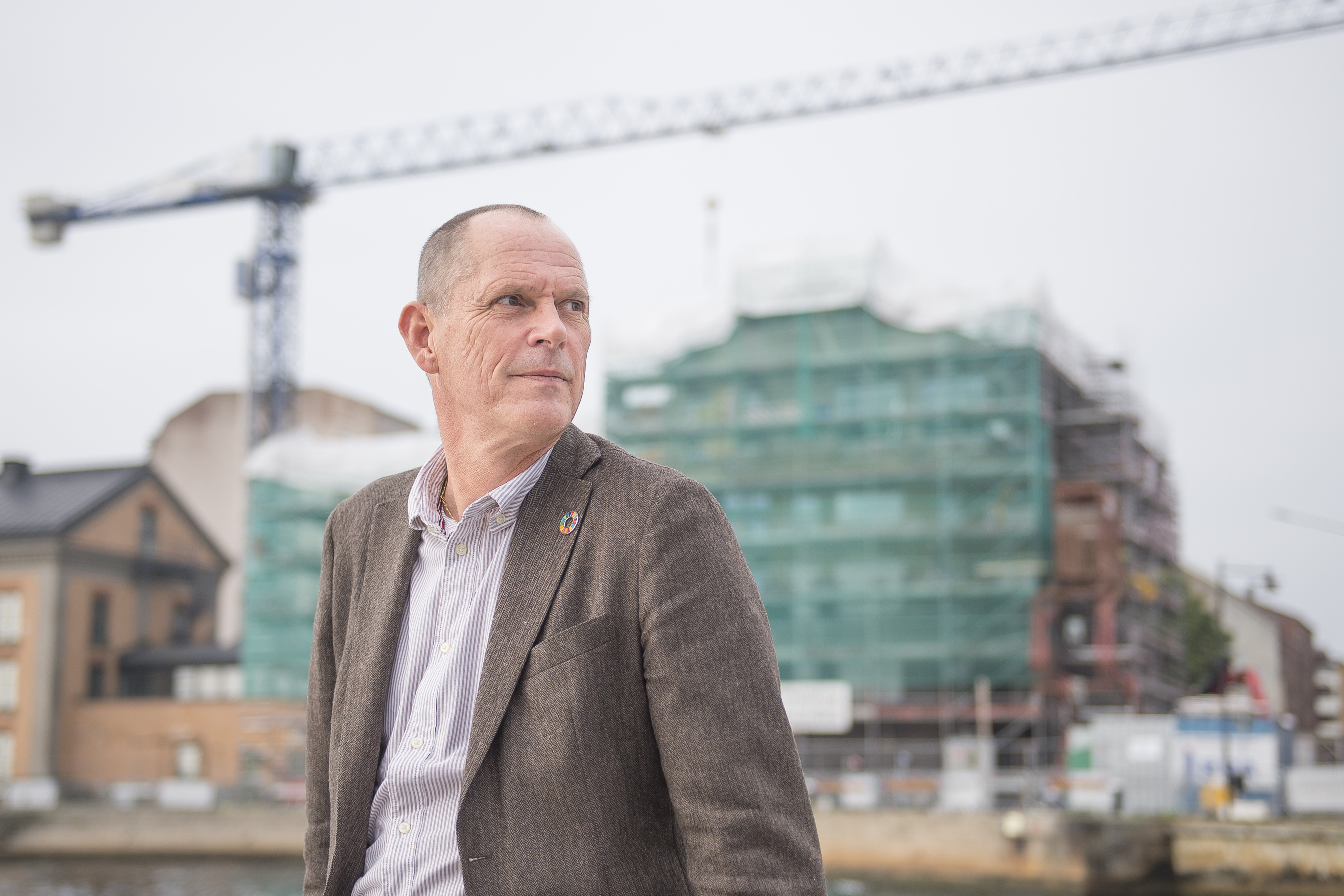 En man i brun kavaj framför en byggarbetsplats med en lyftkran. 