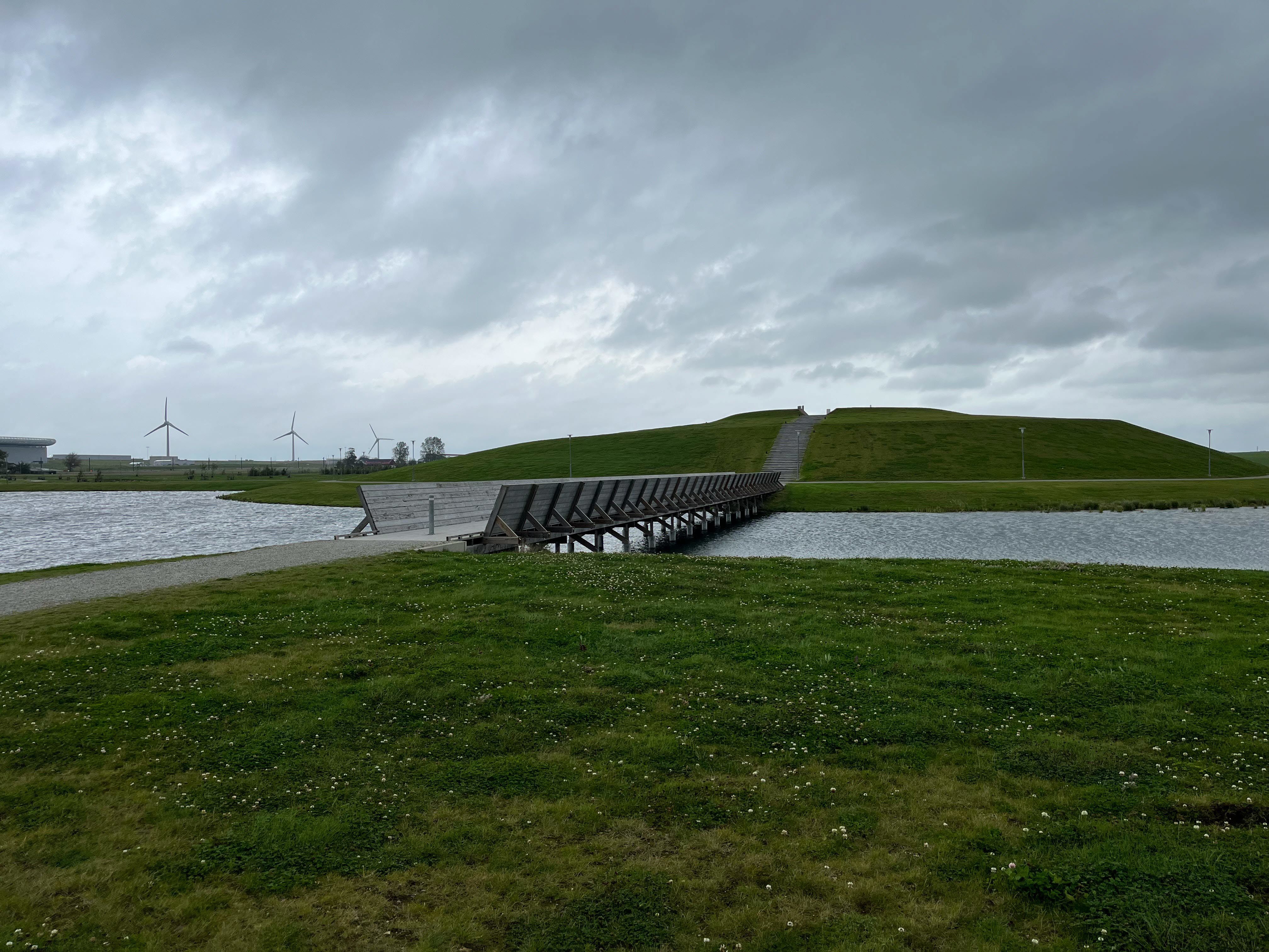 Dagvattendamm med en bro över till en kulle i ett öppet landskap.