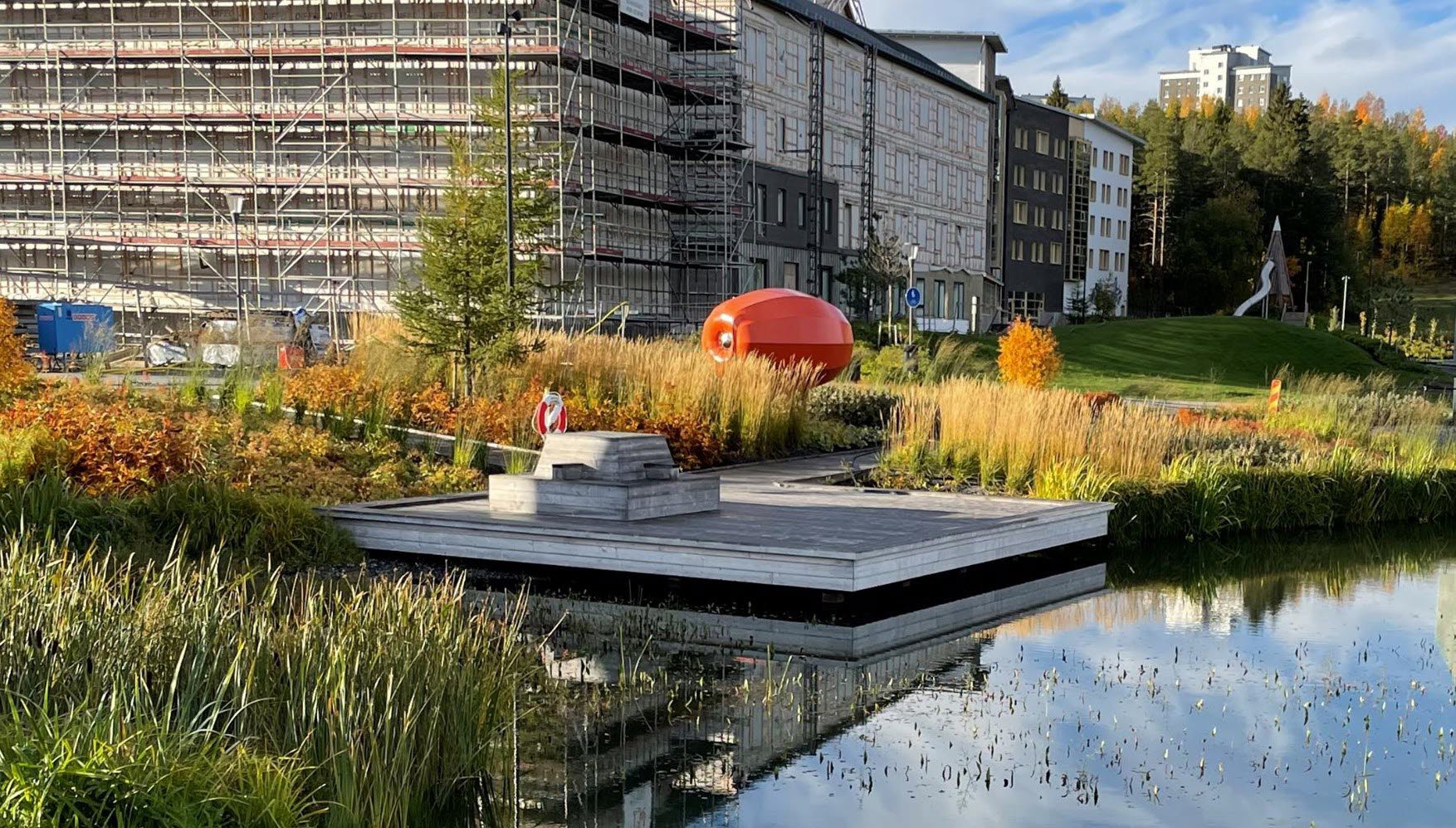 Dagvattendamm med en brygga, byggnader och ett konstverk som ser ut som en stor pumpa.