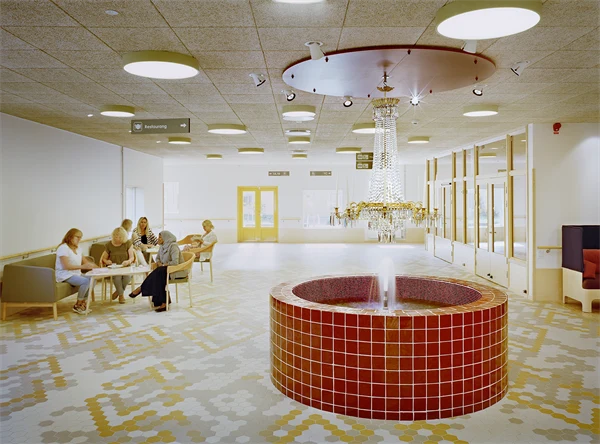 Väntrum där patienter sitter och väntar. I förgrunden finns en rund röd fontän som det hänger en kristallkrona över.