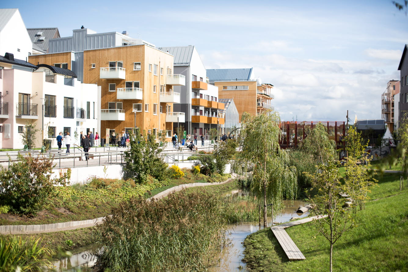 Grönområde med promenadgång utmed slingrande bäck i stadsbebyggelse. Sluttningar med gräs, buskar och träd.