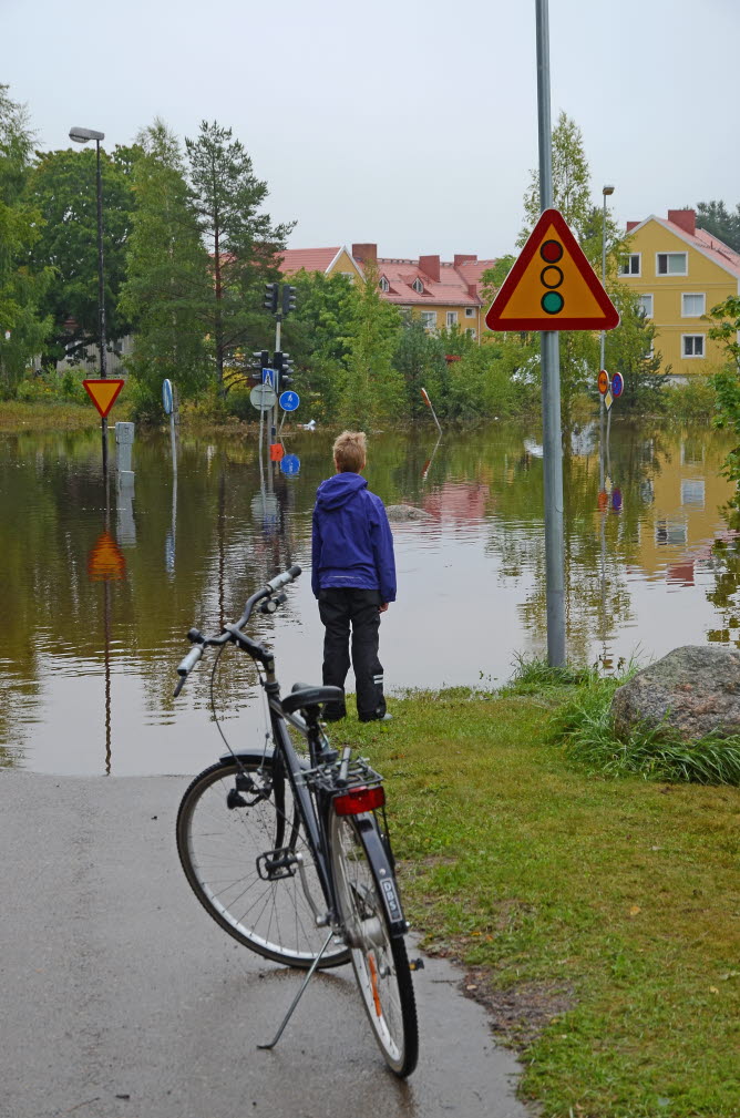 Ett barn har parkerat sin cykel vid en översvämmad vägkorsning. Det går inte att ta sig över till fots eller på cykel.