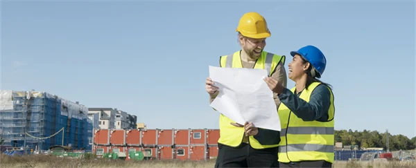 Två personer i varselväst och skyddshjälm pratar och tittar på en ritning framför en byggarbetsplats.