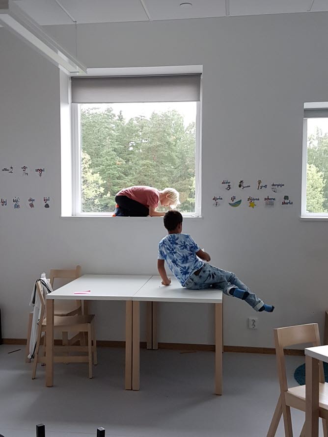 Två barn i ett klassrum. Det ena barnet sitter på ett bord och det andra är hopkrupet i fönsternischen.