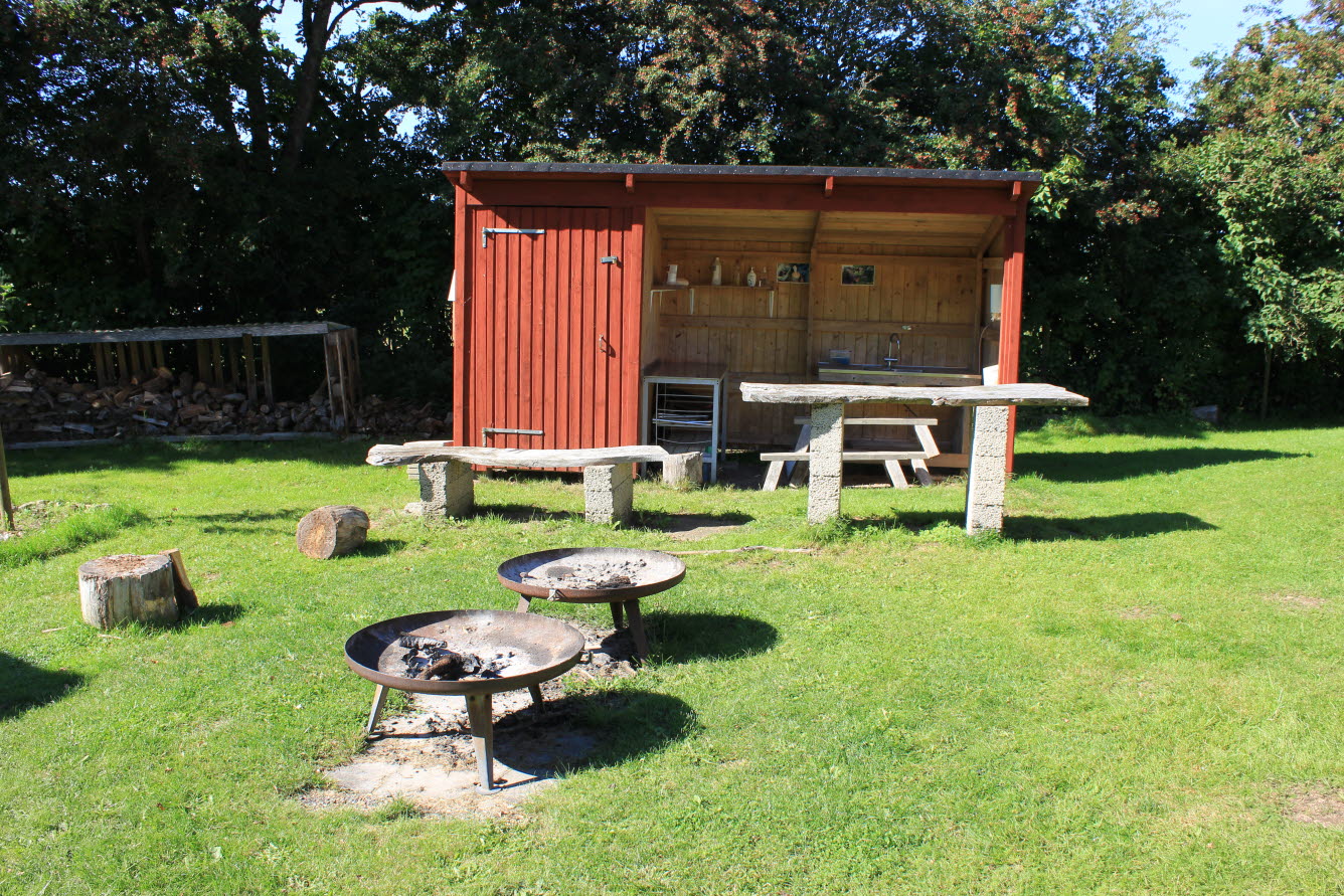 Eldstäder som står på sandbädd i en gräsmatta. Bakom finns bänkar och ett rött skjul med arbetsyta under tak.