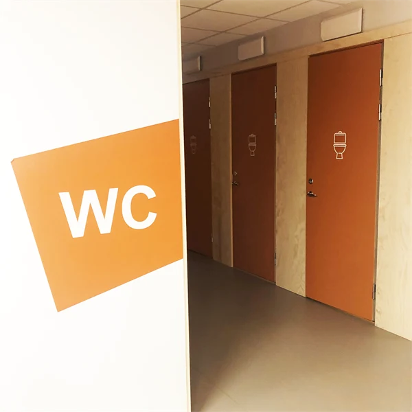 Toalettdörrar med en toalettsymbol. På väggen framför står WC på en orange färgplatta.
