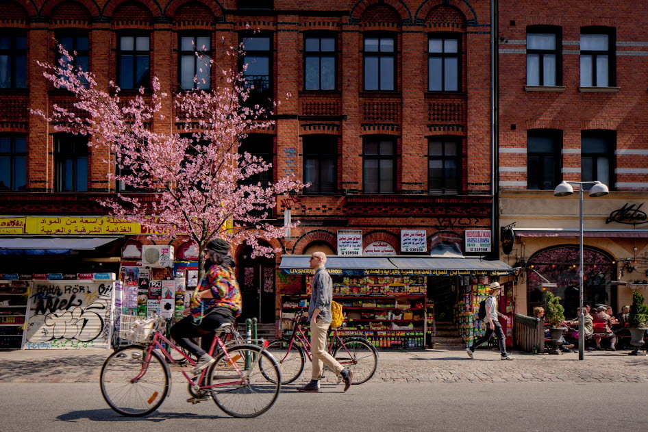 Bild på stadsmiljö i Malmö. Två cyklister på en gata med folkliv och butiker.
