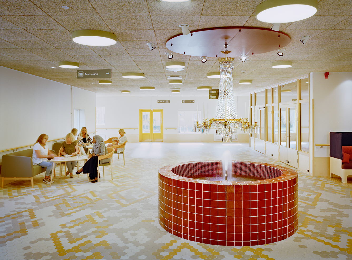Väntrum där patienter sitter och väntar. I förgrunden finns en rund röd fontän som det hänger en kristallkrona över.
