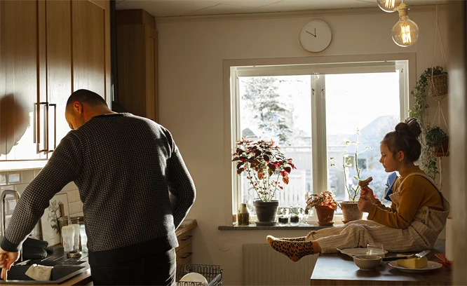 Pappa och flicka i ett kök. Pappan sätter in disk i diskmaskinen och flickan äter smörgås.