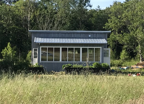 Ett litet grått hus som står bakom en äng före ett skogsparti. Fönsterpartier vetter ut mot ängen.