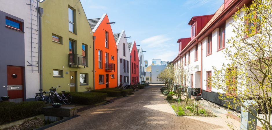 En gata med marksten, på sidorna finns bostadshus i olika färger. Solen skiner.