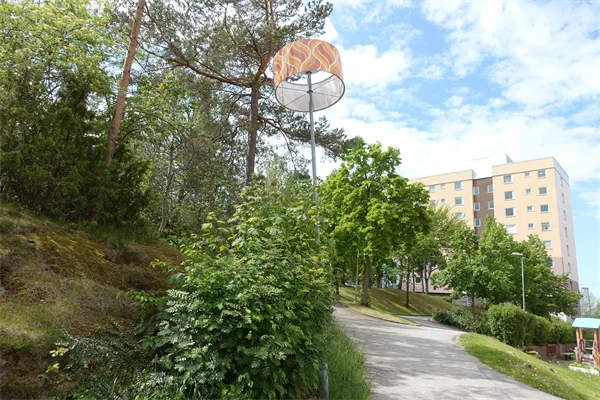 Lampa med lampskärm i park med flerbostadshus i bakgrunden.