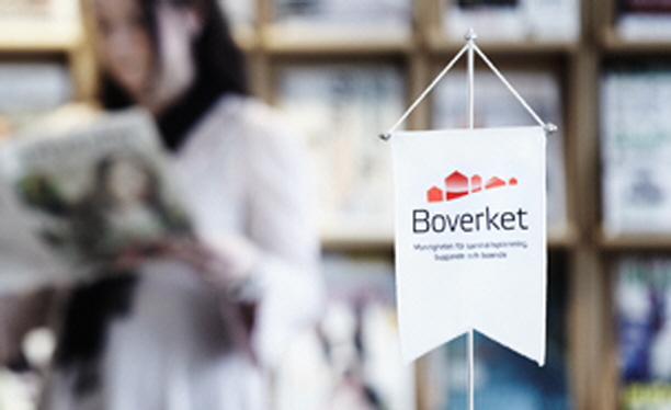 Foto på en bordsvimpel med Boverkets logotyp. En kvinna i bakgrunden som läser i en tidskrift. Foto: Morgan Karlsson. 