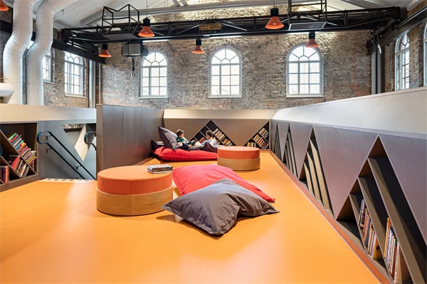 Bibliotek med triangelformade bokhyllor och ett orange golv där det finns orangea sittpuffar.