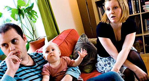 En man sitter i en soffa med ett litet barn i famnen och en kvinna sitter bredvid