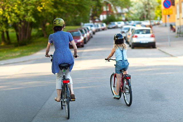 En vuxen och ett barn cyklar på en väg med parkerade bilar.