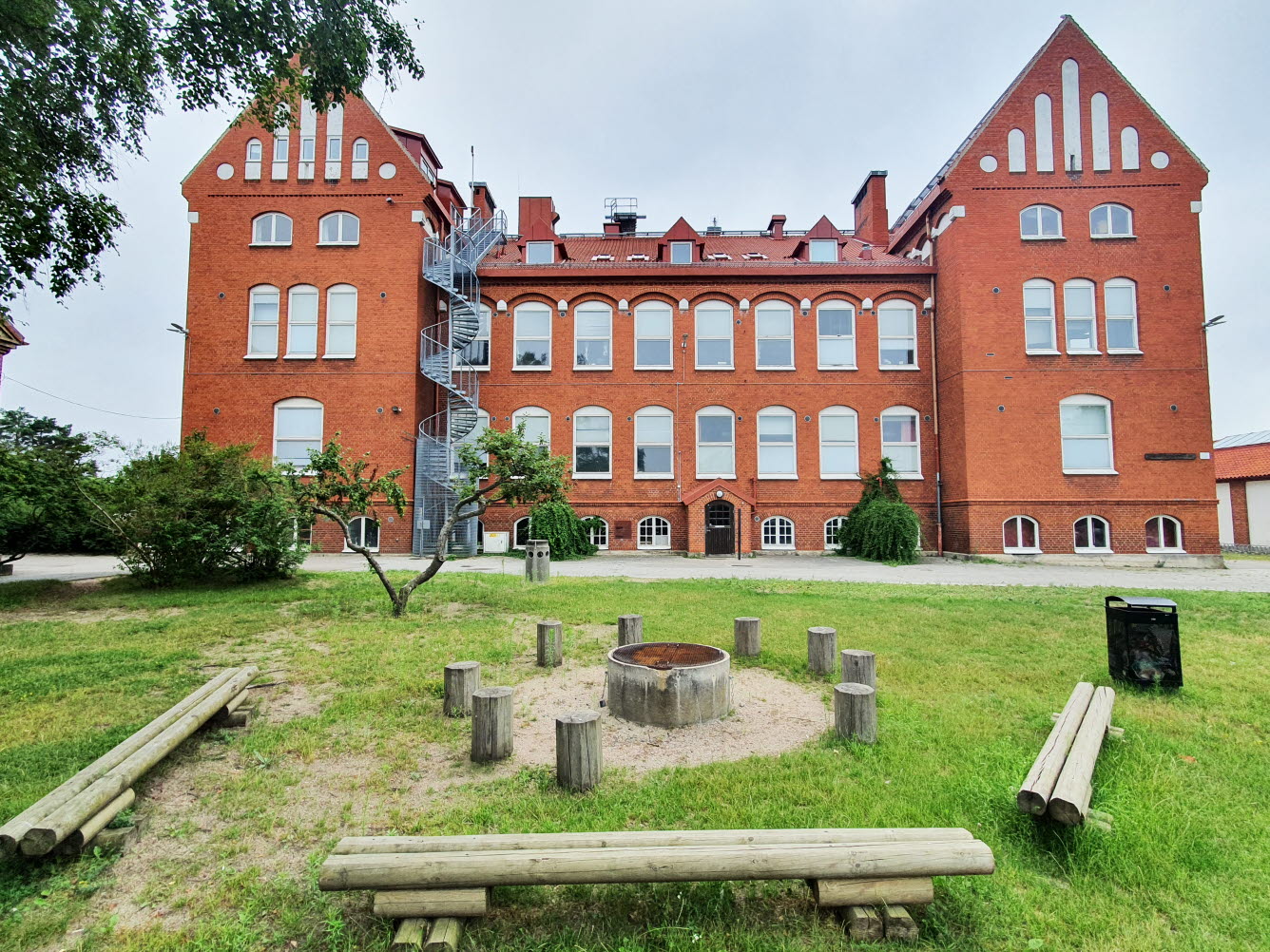 Sittbänkar och sittplatser av stockar. I mitten finns en rund grillplats. I bakgrunden den stora röda skolbyggnaden.