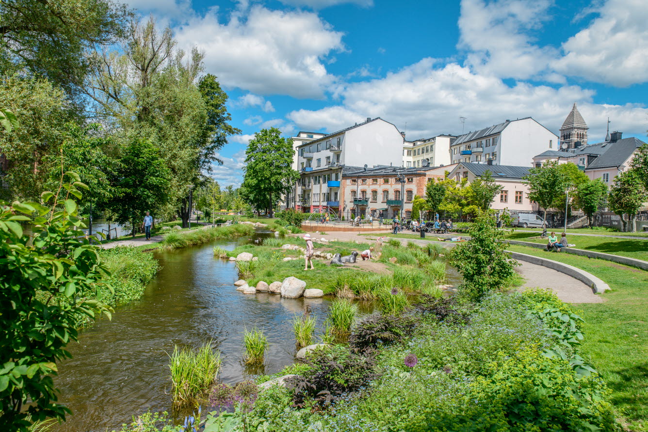 Grönskande parkmiljö med slingrande bäck, promenadvägar, sittplatser och lekpark intill blandad stadsbebyggelse. 