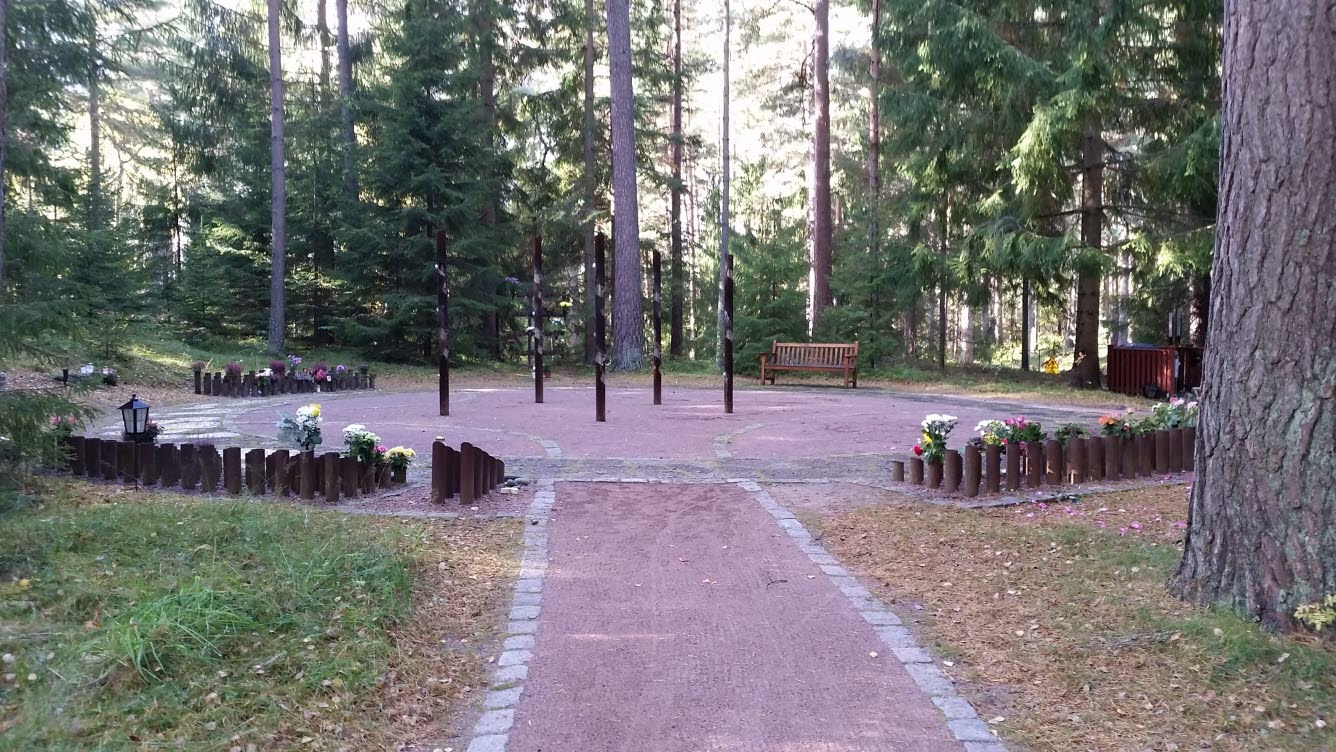 Foto av en minneslund i en skogsglänta. Grusad väg leder fram till en rund yta med blomvaser runt om.