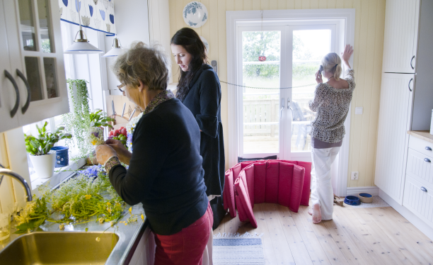 Tre kvinnor i ett kök. Två av dem står vid diskbänken och en står vid altandörren och talar i telefon. Foto: Franz Feldmanis