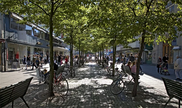 Skuggande trädrad på solbelyst lågfartsgata med  människor.