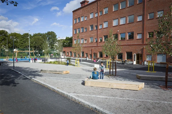 På marken ligger grus och det finns bänkar och små träd på skolgården. I bakgrunden står gungorna.
