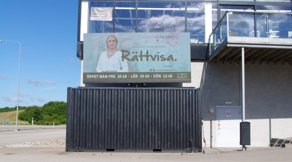 Foto på en stor bildskärm med reklam för Kvinnojouren, ståendes på en svart container vid en affär.