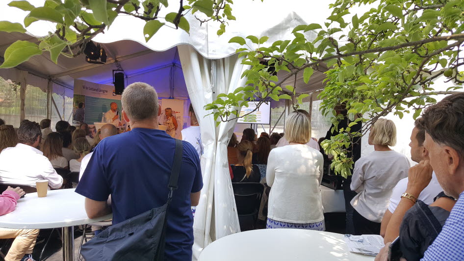 Personer i publik står runt bord och tittar från kameran mot en scen.