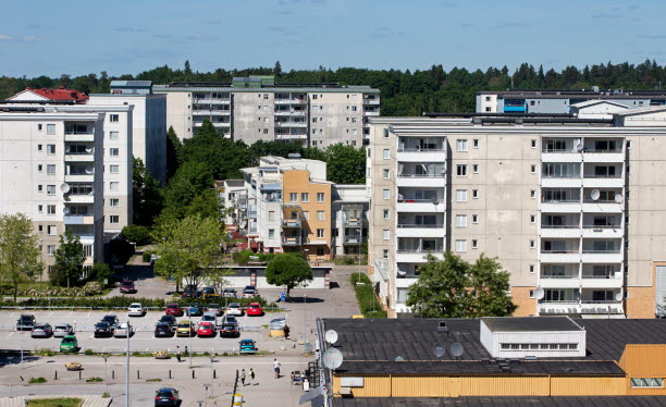 Fyra flerbostadshus och en parkeringsplats med bilar och människor.