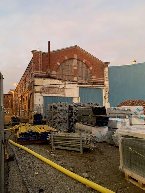 Foto av diverse byggmaterial med en äldre industribyggnad i tegel bakom.