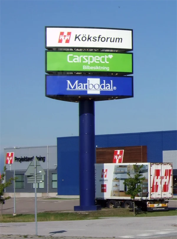 Foto av en hög stolpe med tre skyltar med texten Köksforum, Carspect Bilbesiktning och , Marbodal vid ett köpcentrum.