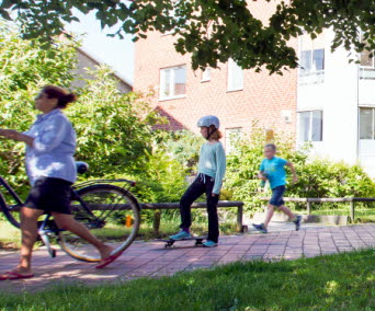 Cyklist, barn på skateboard och joggare på en trottoar framför ett flerbostadshus i en lummig miljö. Det är sommar.
