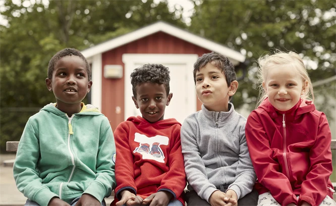 Foto med fyra barn som sitter på rad framför en faluröd bod.